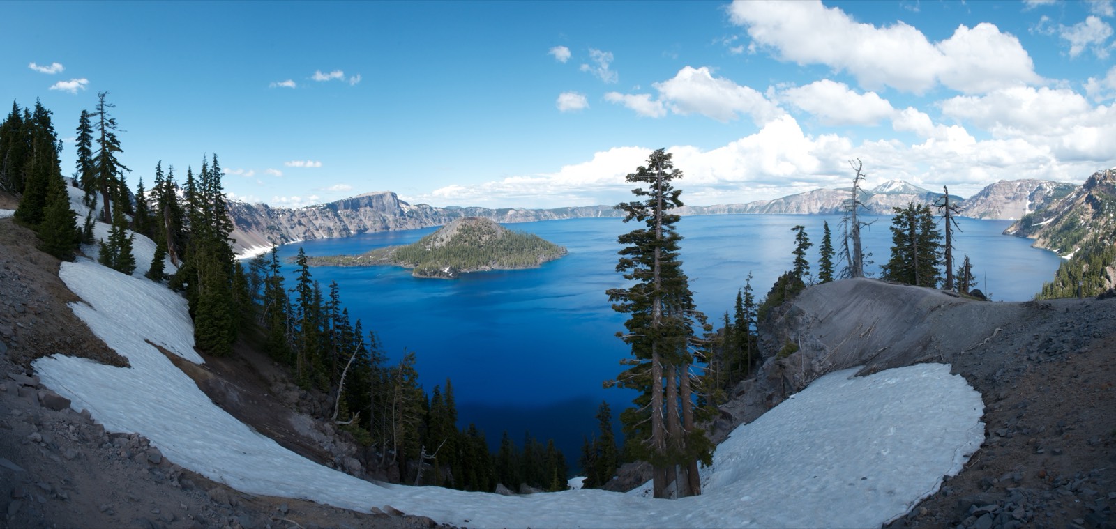 Panoramic view of Crater Lake's deep blue water ringed by caldera cliffs, with the forested cone of Wizard Island rising from the lake and patches of snow on the rim