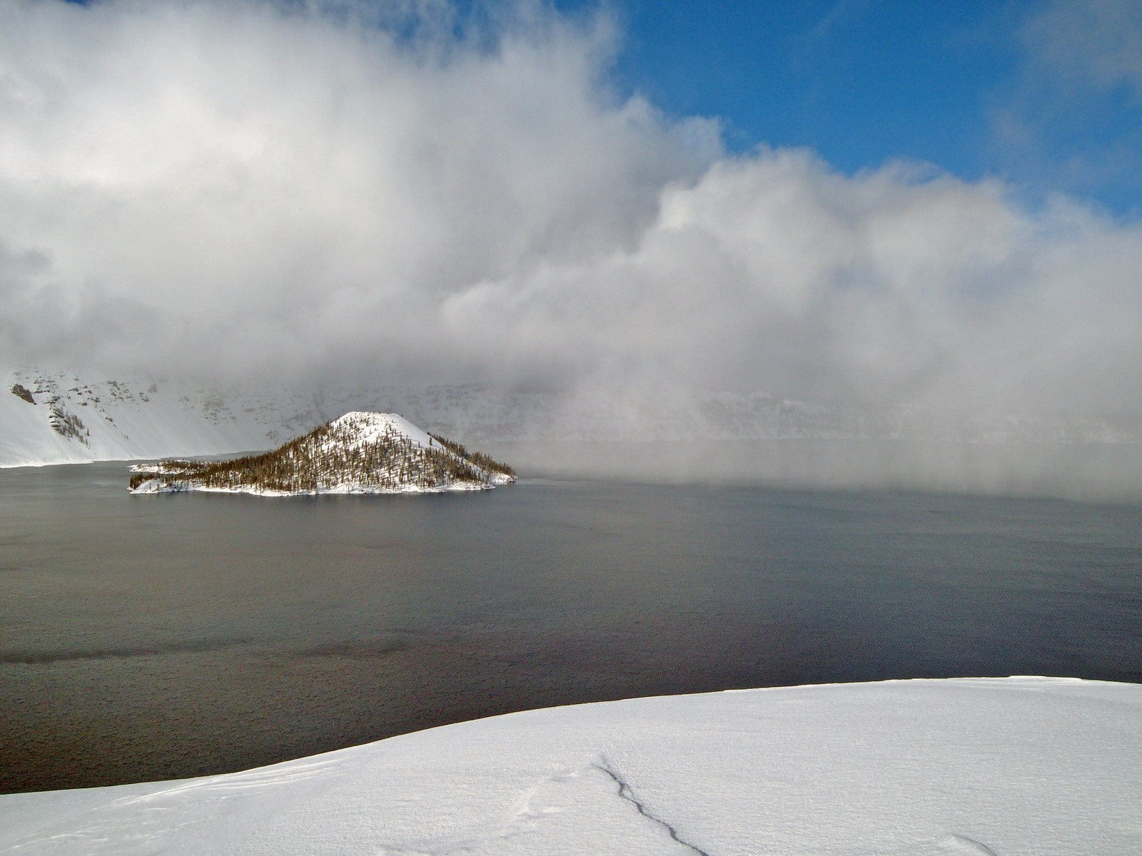 A wall of snow squall cloud sweeping across the surface of Crater Lake in winter, the snow-covered cone of Wizard Island standing in the gray water