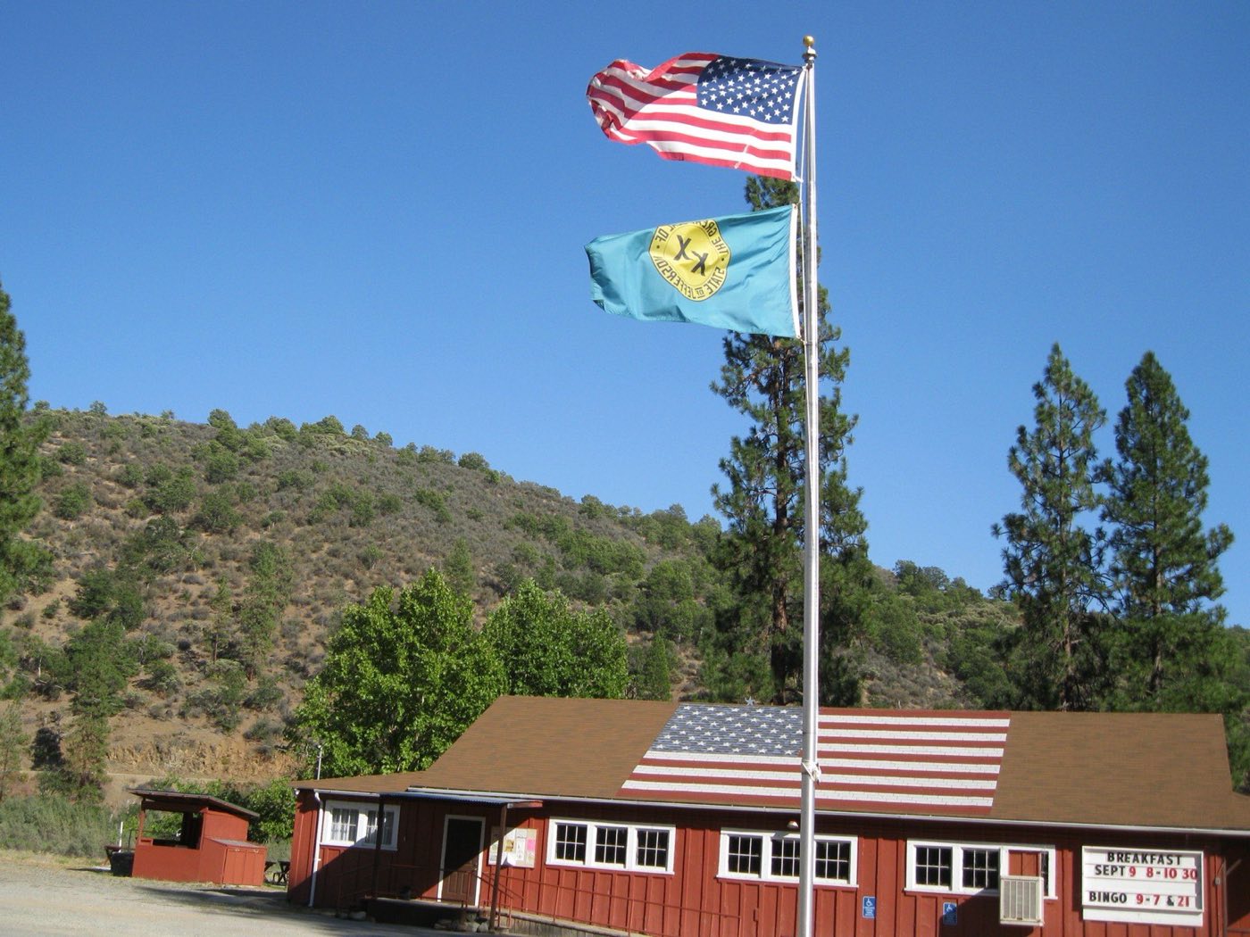 Green State of Jefferson flag with the gold double-cross seal flying under an American flag on a pole in front of a red hall with a US flag painted on its roof