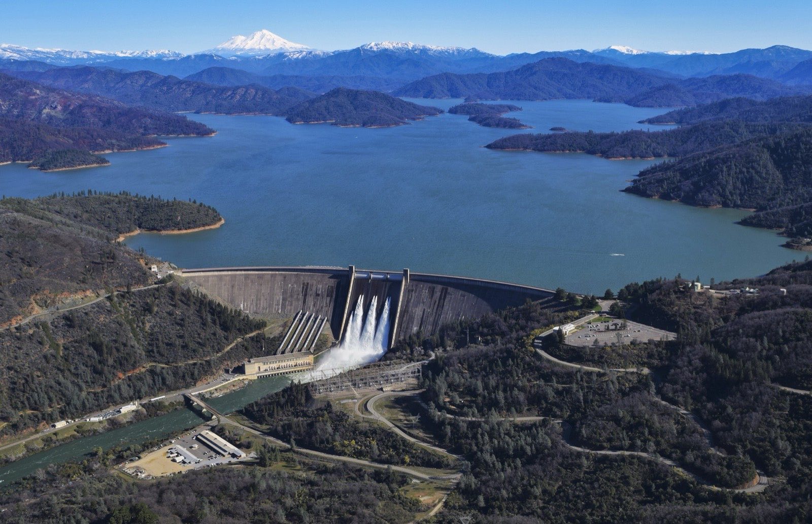 Aerial view of Shasta Dam spilling water into the Sacramento River, with Shasta Lake stretching behind it and Mount Shasta on the horizon